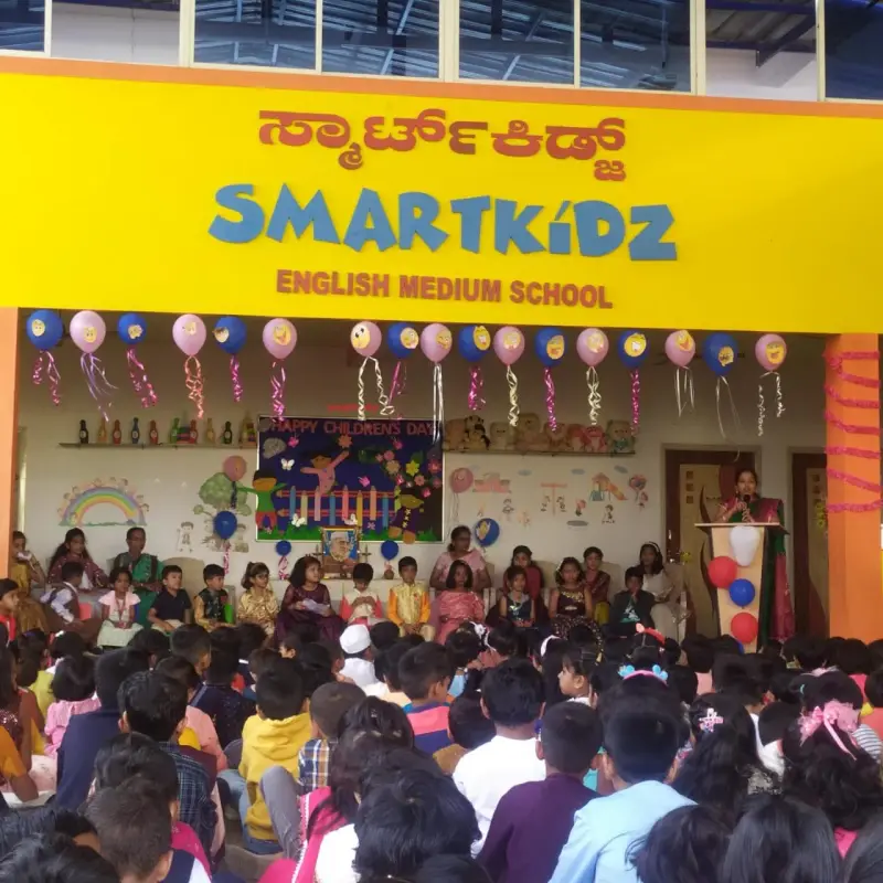 Children sit facing a stage decorated for Happy Children's Day under a SMARTKiDZ English Medium School sign, while a woman speaks at a lectern.