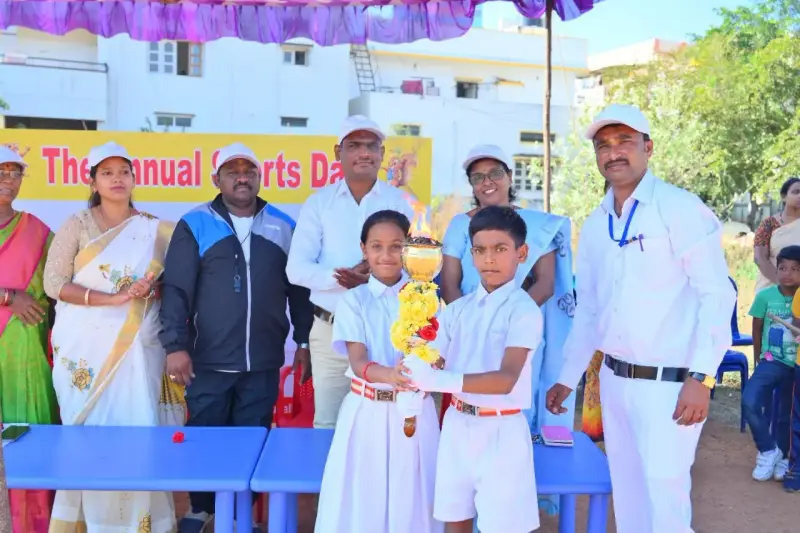 Two children in white uniforms hold a decorated torch in front of adults, beneath a banner that reads The annual Sports Day.